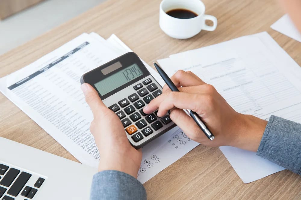 A pair of hands working a calculator over printed ledgers, a laptop and a coffee on the desk